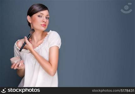 Portrait of a Beautiful woman with makeup brush near her face looking at camera and smiling against grey background