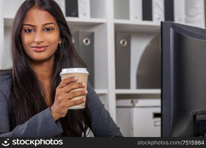 Portrait of a beautiful young Asian Indian woman or businesswoman in office using a computer and drinking takeout tea or coffee