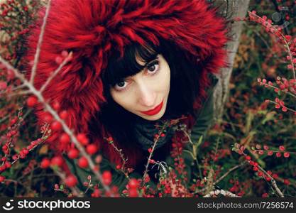 Portrait of a beautiful young brunette woman in red with a smile and hood with red hair in the forest behind a bush with red berries.
