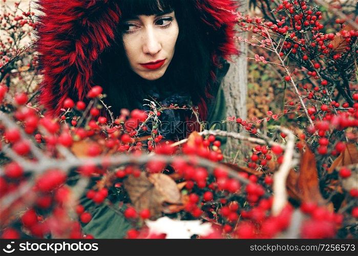 Portrait of a beautiful young brunette woman in red with hood with red hair in the forest behind a bush with red berries.