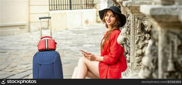 Portrait of a beautiful young caucasian traveling woman sitting on the street with suitcase smiling and looking at the camera outdoors. Traveling woman sitting at the door