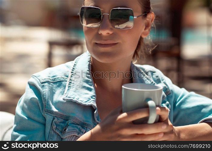 Portrait of a Beautiful Young Female Wearing Stylish Sunglasses Sitting in Outdoors Cafe with Cup of Coffee in Hands. Enjoying Sunny Summer Day in the City. Coffee Break