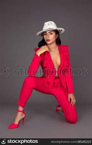 Portrait of a beautiful young white woman with wavy black hair and beautiful makeup posing by herself inside a studio with a grey background wearing a red business suit with high heels and a white hat.