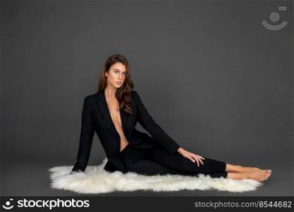 Portrait of a beautiful young white woman with wavy hair and beautiful makeup posing by herself laying on fur inside a studio with a grey background wearing a black business suit.