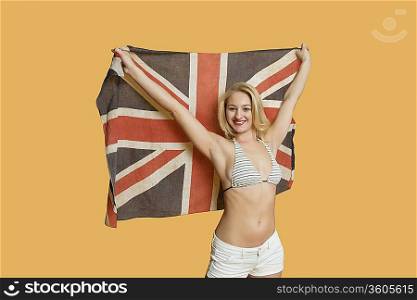Portrait of a beautiful young woman holding British flag with arms raised over colored background
