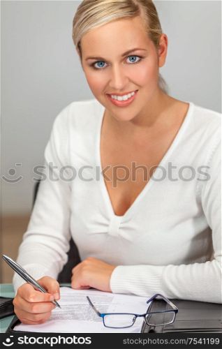Portrait of a beautiful young woman or businesswoman with perfect white teeth working at her desk in an office writing and smiling to camera