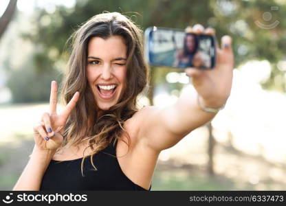 Portrait of a beautiful young woman selfie in the park with a smartphone doing v sign