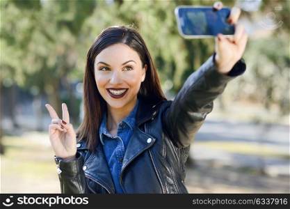 Portrait of a beautiful young woman selfie in the park with a smart phone. Girl doing V sign.