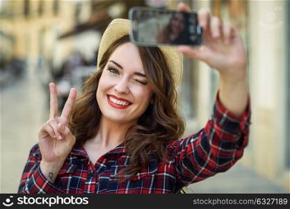 Portrait of a beautiful young woman selfie in the street with a smartphone. Young woman wearing plaid shirt and sun hat with red lips in urban background.