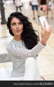 Portrait of a beautiful young woman selfie in urban street with a smartphone. Brunette girl with long dark hair blowing a kiss.