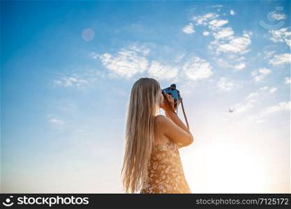 portrait of a blonde girl in a floral print dress with a vintage video camera in a grape field records video of a plane taking off