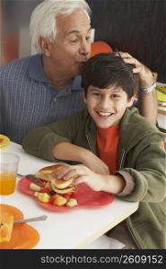 Portrait of a boy holding a burger with his grandfather kissing him on his head