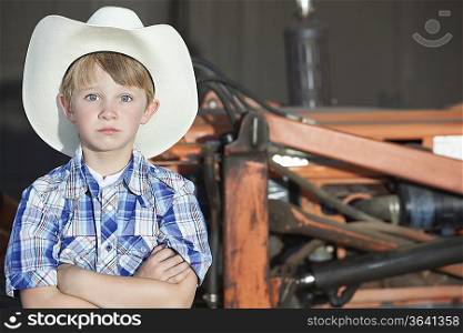 Portrait of a boy wearing cowboy hat while standing with arms crossed against machine