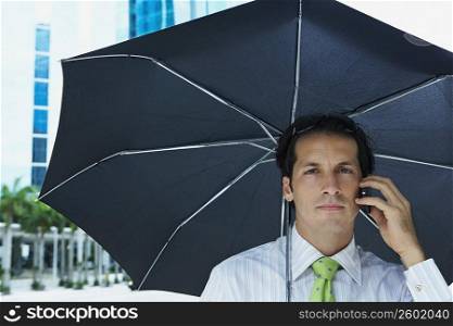 Portrait of a businessman talking on a mobile phone under an umbrella