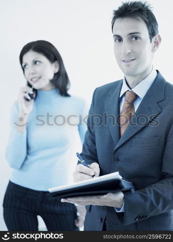 Portrait of a businessman writing on a personal organizer with a businesswoman standing behind him holding a mobile phone