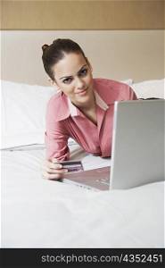 Portrait of a businesswoman holding a credit card and lying in front of a laptop on the bed
