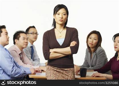 Portrait of a businesswoman leaning against a table with business executives sitting behind her