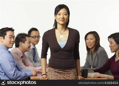 Portrait of a businesswoman leaning against a table with business executives sitting behind her