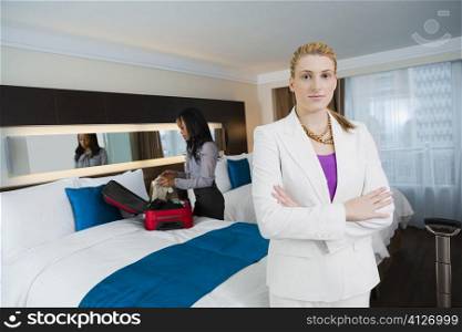 Portrait of a businesswoman standing with her arms crossed with another businesswoman packing her luggage