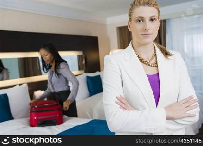 Portrait of a businesswoman standing with her arms crossed with another businesswoman packing her luggage