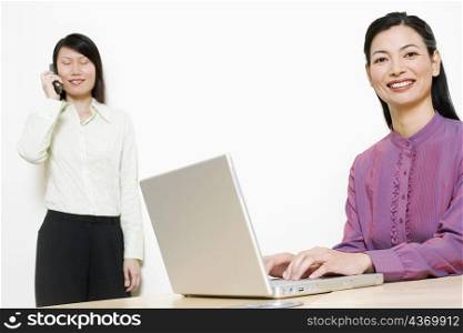 Portrait of a businesswoman using a laptop with another talking on mobile phone behind her