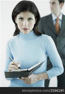 Portrait of a businesswoman writing on a personal organizer with a businessman standing behind her
