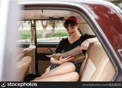 portrait of a Caucasian beautiful young girl in a black vintage dress, posing in the salon of vintage car reading an old magazine