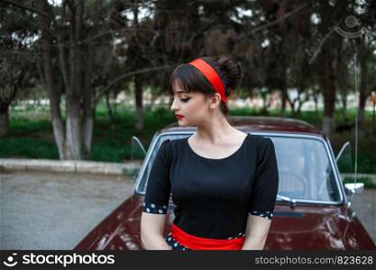 portrait of a Caucasian beautiful young girl in a black vintage dress, posing near a vintage car