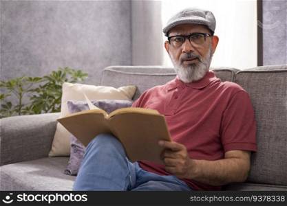 Portrait of a cheerful senior man with flat cap reading a book while sitting on sofa at home