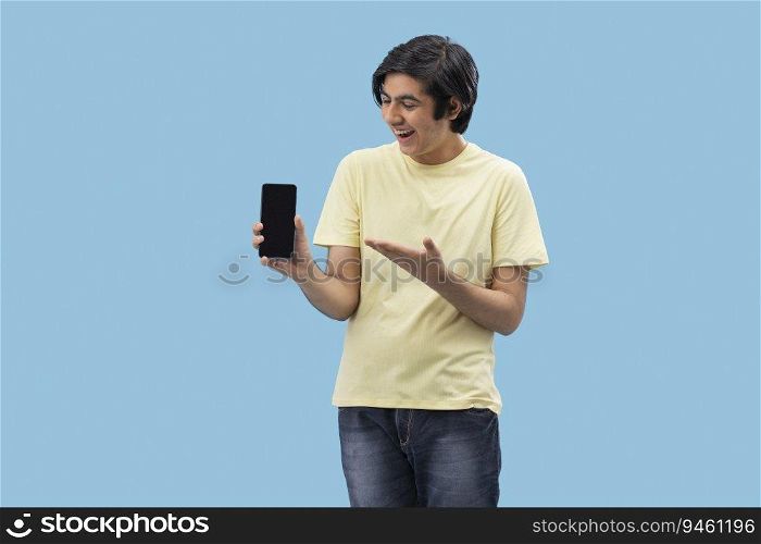 Portrait of a cheerful teenage boy holding a Smartphone and gesturing against blue background