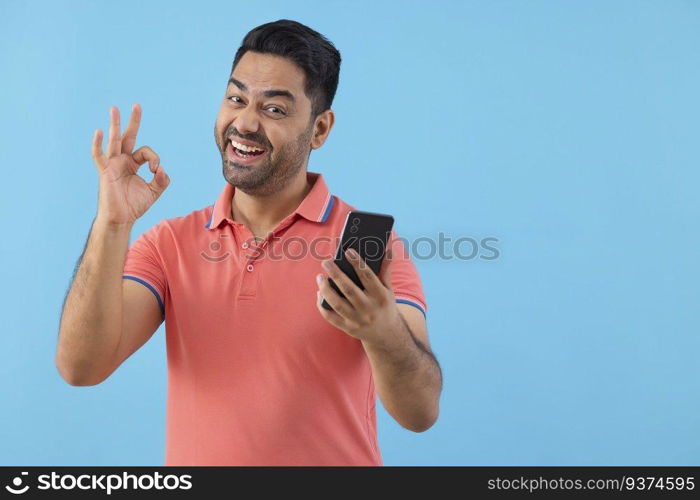 Portrait of a cheerful young man holding Smartphone and showing OK sign