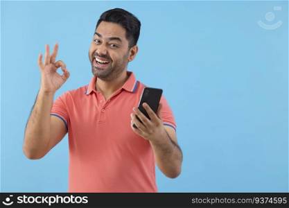 Portrait of a cheerful young man holding Smartphone and showing OK sign