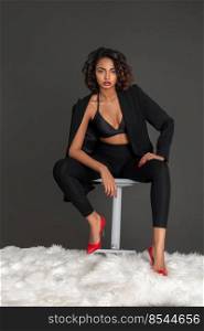 Portrait of a confident black woman with curly dark hair and beautiful makeup sitting by herself on a bed of fur inside a studio with a grey background wearing a black jacket with red high heels.