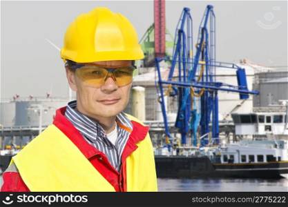 Portrait of a confident docker foreman in front of a petrochemical harbor with gas installations, numerous cranes, and a freight ship