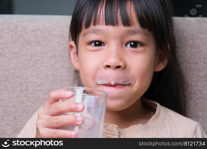 Portrait of a cute Asian little girl holding a glass of milk sitting on the sofa at home. Small girl at home with smiling face, feeling happy enjoying drinking milk and looking at camera.