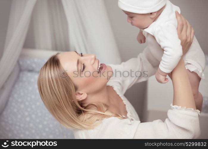 Portrait of a cute blond young mother playing with her adorable baby at home, happy mom lifting up her little son, enjoying parenthood