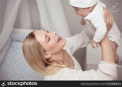 Portrait of a cute blond young mother playing with her adorable baby at home, happy mom lifting up her little son, enjoying parenthood
