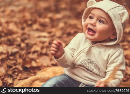 Portrait of a cute child spending time in the autumn forest, with pleasure sitting on the blanket on the ground covered with dry tree leaves and smiling, happy carefree childhood
