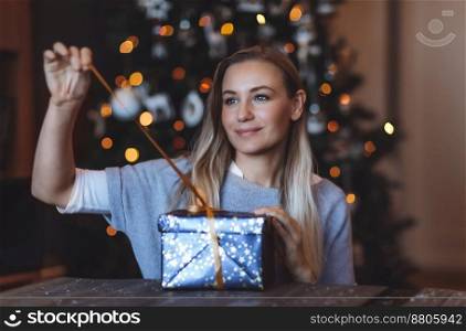 Portrait of a Cute Girl opening Festive Gift Box at Home over Decorated Christmas Tree Background. Happy Winter Holidays. Enjoying Celebration.. Beatiful Woman Opens Present