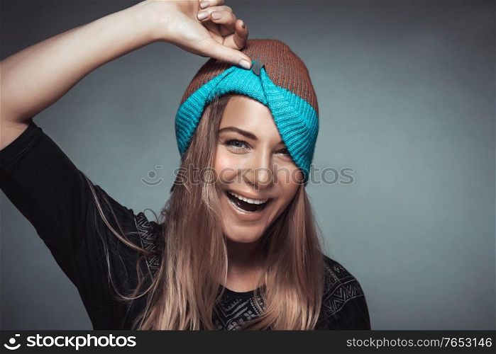 Portrait of a cute happy cheerful girl making faces and playing with a hat. Young model over gray background, funky urban style fashion for youngsters, joyful life