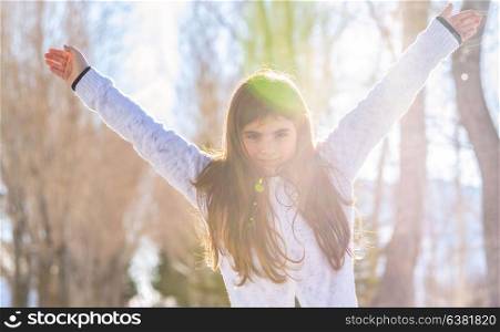 Portrait of a cute happy little girl with raised up hands in the forest enjoying bright sunny winter day, having fun outdoors, good warm weather in the wintertime
