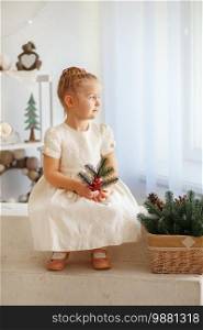 Portrait of a cute little blonde girl holding fir twig and looking out the window in the interior with Christmas decorations. Beautiful Little girl waiting for a miracle in Christmas decorations
