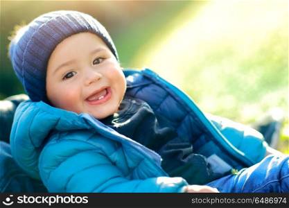 Portrait of a cute little boy enjoying spring sunny day, wearing blue jacket and hat sitting on the grass field, happy day outdoors. Happy day of a cute boy