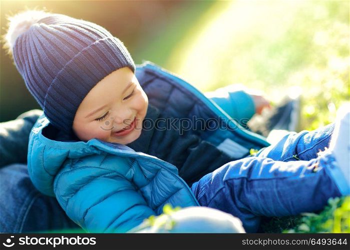 Portrait of a cute little boy wearing blue jacket and hat sitting on green grass field, laughing and having fun outdoors, small baby enjoying spring sunny day. Joyful baby time