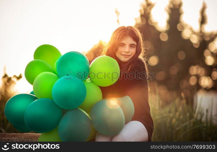 Portrait of a cute little girl sitting in the park with pile of green air balloons outdoors in mild evening sun light, celebrating birthday, preparation to happy holiday