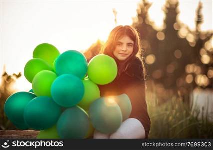 Portrait of a cute little girl sitting in the park with pile of green air balloons outdoors in mild evening sun light, celebrating birthday, preparation to happy holiday