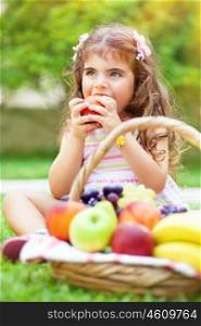 Portrait of a cute little girl sitting on the green grass field in a park and with pleasure eating tasty juicy apple, healthy babies nutrition