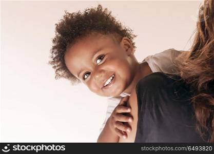 Portrait of a cute little smiling baby looks out from behind the mother&rsquo;s shoulder, happy african american family isolated on beige white background, love and togetherness concept. Cute little boy with mother