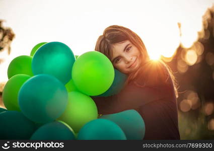 Portrait of a cute smiling girl outdoors hugging big pile of green air balloons, preparing to celebrate birthday party, happy carefree childhood 