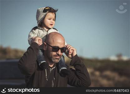 Portrait of a father carrying his cute little son on shoulders, baby and dad with pleasure spending time together outdoors, happy family life. Father playing with his little son